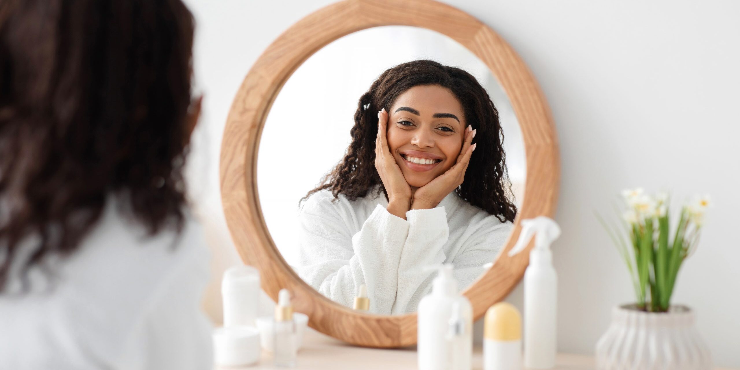Great result, shiny clean skin, happy morning and perfect beauty. Smiling young african american woman in white coat touches face and looks in round mirror at table with cosmetics in bedroom interior