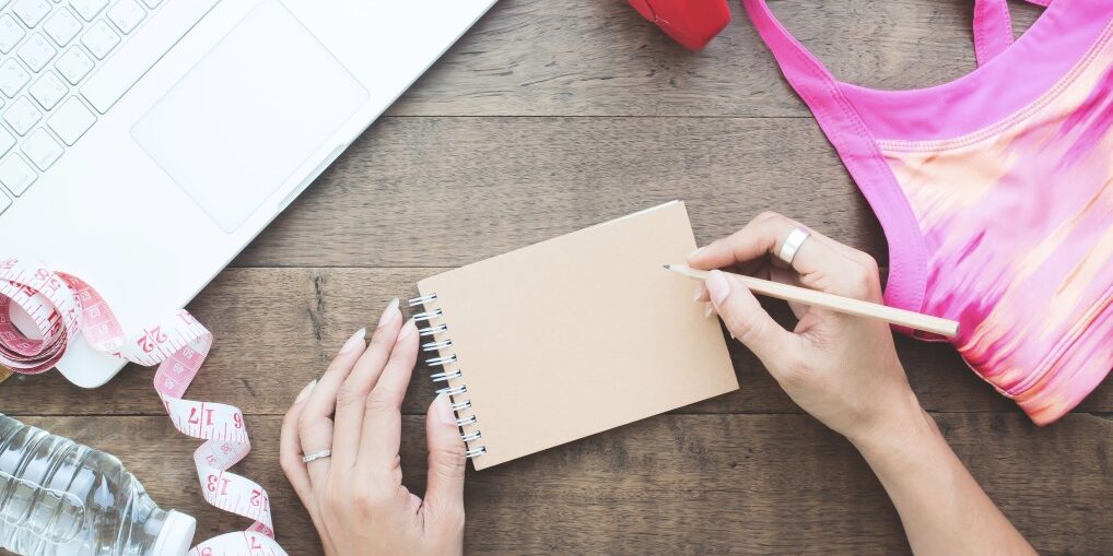 overhead-view-of-woman-hand-writing-on-notebook-with-laptop-and-fitness-items-on-wooden-table_t20_OpVZdL