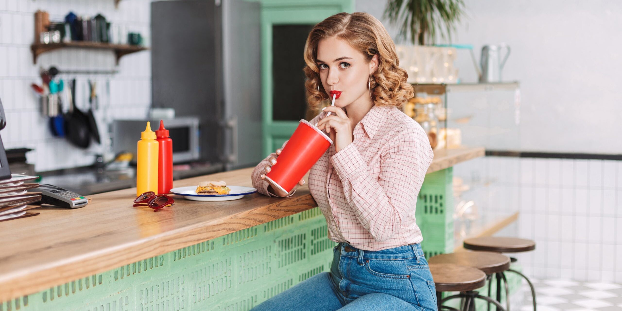 Pretty girl in shirt and jeans sitting at the bar counter in cafe and drinking soda water while dremily looking in camera